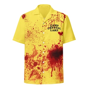 May include: Yellow short-sleeved button-down shirt with a camp collar. The shirt features a splatter design in red and the text "CAMP CRYSTAL LAKE" in black.