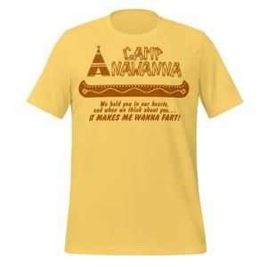 Op de afbeelding: Mosterdgeel T-shirt met de tekst "CAMP NAWANNA" boven een kano-afbeelding. Onder de kano staat de tekst: "We hold you in our hearts, and when we think about you... IT MAKES ME WANNA FART!"