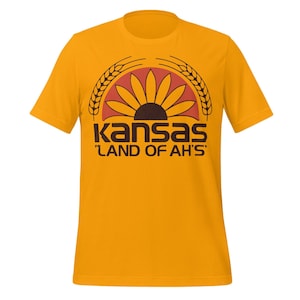 Może przedstawiać: Musztardowa koszulka z grafiką słonecznika, kłosów pszenicy i napisem "Kansas" w kolorze czarnym, z tekstem "LAND OF AH'S" poniżej. Projekt jest w odcieniach czerwieni, brązu i żółci.