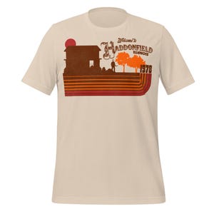Könnte beinhalten: Cremefarbenes T-Shirt mit Retro-Grafik. Das Design zeigt eine Silhouette eines Hauses, einer Figur und Bäume, mit dem Text "Welcome to Haddonfield Illinois 1978." Die Grafik verwendet Orange-, Braun- und Rottöne.