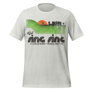 May include: A light grey t-shirt with a graphic design. The design features the text "Visit Sing Sing Correctional Facility • Ossining, New York." It includes a green hill, orange sun, and a black building silhouette.