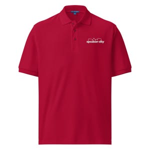 May include: A red polo shirt with short sleeves and a collared neckline. The shirt features a three-button placket and a white logo that reads "speaker city" on the upper left chest.