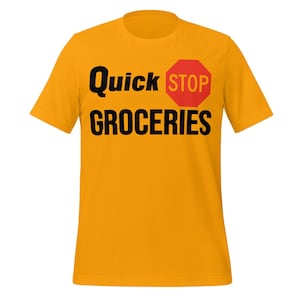 Puede incluir: Camiseta de color amarillo mostaza con las palabras "Quick Stop GROCERIES" en negro, con un gráfico de señal de stop roja. La camiseta es de cuello redondo y manga corta.