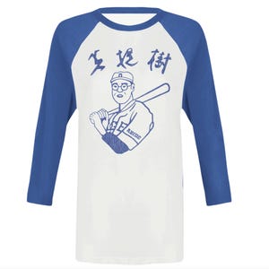 Op de afbeelding: Een wit en blauw baseball-shirt met een cartoon illustratie van een honkbalspeler die een knuppel vasthoudt. Het shirt heeft blauwe mouwen en een witte body met Japanse tekst en het woord "ABIDE" op de voorkant.