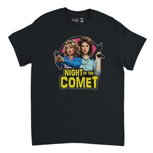 Puede incluir: Camiseta negra con un diseño gráfico de la película "Night of the Comet". El diseño muestra a dos mujeres sosteniendo armas, sobre un fondo rosa y amarillo. El texto "NIGHT OF THE COMET" se muestra de forma destacada.