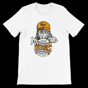 Puede incluir: Camiseta blanca con un diseño temático de béisbol. El diseño incluye una bruja con una gorra de béisbol con "SW" y las palabras "Salem Witches Baseball Club" en una pelota de béisbol. Cuello redondo.