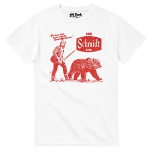 May include: White t-shirt with a red graphic featuring a man with a rifle and a bear, with the text "Schmidt BEER". The shirt also has the quote "The beer that grew with the Great Northwest."