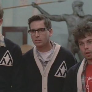 Op de afbeelding: Drie jonge mannen dragen zwarte vesten met witte bies en een wit driehoeklogo. De mannen dragen een bril en overhemden met knopen. E&eacute;n man draagt een rood t-shirt onder een geruit overhemd.