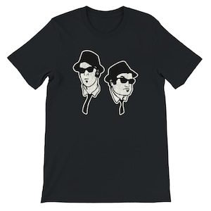 Puede incluir: Camiseta negra con un gráfico de dos hombres con sombreros y gafas de sol. La ilustración tiene un aspecto vintage y texturizado con contornos blancos. Las figuras llevan corbatas y camisas con cuello.