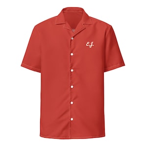 May include: A short-sleeved, red button-down shirt with a classic collar. The shirt features white buttons and the embroidered initials "E.J." on the left chest. The shirt is made of a smooth fabric.