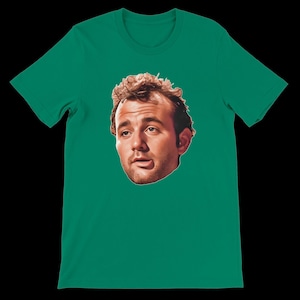 May include: A green t-shirt featuring a portrait of a man's face. The portrait is centered on the shirt and is a close-up of the man's face. The shirt is a solid green color.
