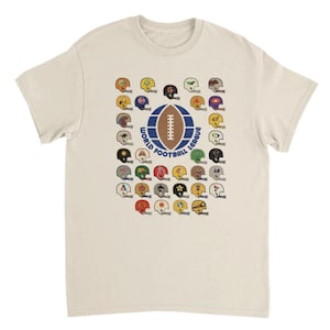 Puede incluir: Camiseta color crema con un gráfico de cascos de fútbol en varios colores y diseños. Un emblema central muestra un balón de fútbol y el texto "WORLD FOOTBALL LEAGUE". La camiseta tiene mangas cortas y cuello redondo.