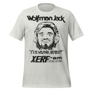 May include: Light gray t-shirt featuring a black and white graphic of Wolfman Jack with the text "Wolfman Jack". Below the image is the text "IT'S DE WOLFMAN, BAYBEEE!!!" and "XERF-am CIUDAD ACUNA".