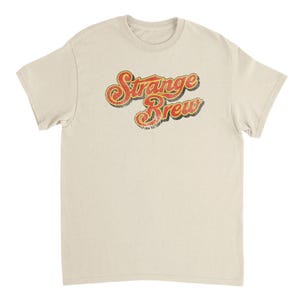 May include: A cream-colored t-shirt with the words "Strange Brew" in a retro, distressed font. The text is in red and yellow with a black outline, giving it a vintage look. The shirt has short sleeves and a crew neck.