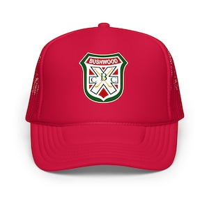 May include: A red trucker hat with a mesh back and a curved brim. The front of the hat features a shield-shaped emblem with the text "BUSHWOOD" and "CBC" in a green, white, and red design. The hat is a classic accessory.