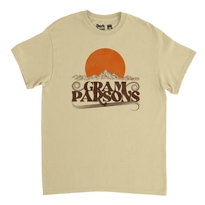 May include: A tan t-shirt featuring a brown graphic of a sunset over mountains with the text "Gram Parsons" in a vintage font. The shirt has a crew neck and short sleeves. The brand logo is visible on the upper back.