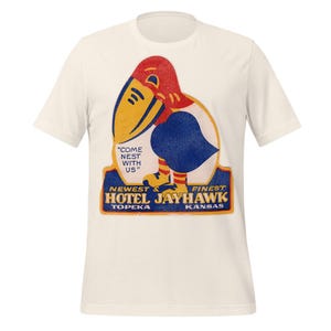 May include: Off-white t-shirt with a retro design of a Jayhawk bird in red, yellow, and blue. The graphic includes the text "COME NEST WITH US" and "HOTEL JAYHAWK TOPEKA KANSAS".