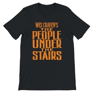 May include: A black short-sleeved t-shirt featuring the orange text WES CRAVEN'S THE PEOPLE UNDER THE STAIRS printed on the front. The lettering has a distressed, weathered appearance with a bold, blocky font style.