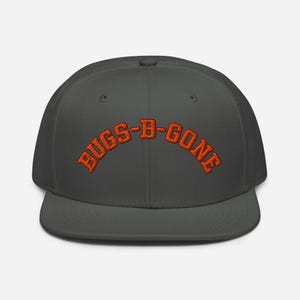 May include: A charcoal gray flat-brimmed baseball cap with the embroidered text "BUGS-B-GONE" in orange. The cap has a structured fit and a flat visor. The text is arched across the front of the cap.