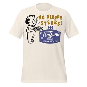 Può includere: T-shirt color crema con un'illustrazione in stile vintage. La grafica include un personaggio dei cartoni animati che tiene un piatto di cibo, con il testo "NO SLOPPY STEAKS!" sopra un logo blu e bianco che dice "Truffoni's Original Steak House."