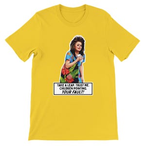 Puede incluir: Camiseta amarilla con un gráfico de una mujer con un vestido floral sosteniendo una taza. El texto en la camiseta dice: "TAKE A LEAP. TRUST ME. CHILDREN POINTING. YOUR FAULT!"