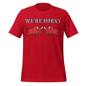 May include: A red t-shirt featuring the text "WE'RE HORNY" above a graphic of two red bulls facing each other. The bulls have white horns and are in a charging pose. The shirt is made of a soft material.