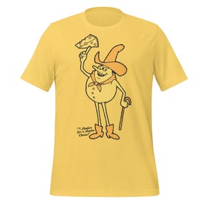 May include: Yellow t-shirt featuring a cartoon character holding a cheese wedge. The character wears a cowboy hat, neckerchief, and boots, and carries a cane. Text on the shirt reads "I Hanker for a Hunk a Cheese!"