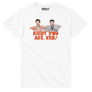 May include: White t-shirt featuring a graphic of two men with their arms outstretched, with the text "RIGHT YOU ARE, KEN!" in orange. The shirt has a crew neck and short sleeves. The brand logo is visible at the top.