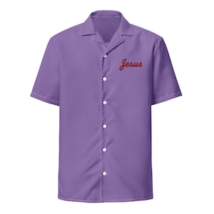 Puede incluir: Camisa abotonada de manga corta en color lavanda liso. La camisa presenta un cuello clásico y la palabra "Jesus" en cursiva roja en el pecho. La camisa tiene botones blancos en la parte delantera.