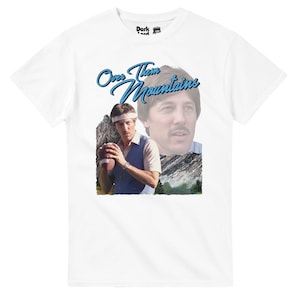 May include: White t-shirt with a retro graphic of a man holding a football, and the blue text "Over Them Mountains". The design includes a mountain backdrop and a second image of the man's face.