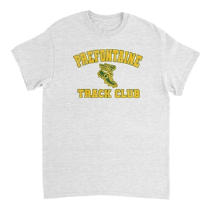 May include: A light gray t-shirt featuring the text "PREFONTAINE TRACK CLUB" in yellow and green. The design includes a running shoe graphic. The shirt is a classic crew neck style.