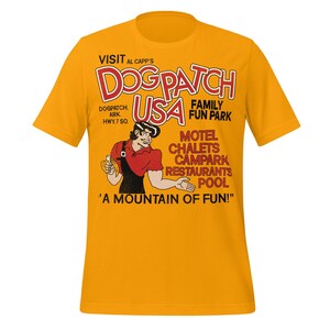 May include: Mustard yellow t-shirt featuring a vintage cartoon graphic. The graphic includes the text "VISIT AL CAPP'S DOGPATCH USA FAMILY FUN PARK" with a cartoon character and additional text. The shirt is a classic crew neck style.