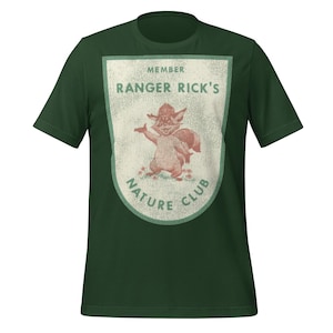 Può includere: T-shirt verde scuro con una grafica invecchiata di una volpe dei cartoni animati con un cappello e la scritta "MEMBER RANGER RICK'S NATURE CLUB". Il design è nei colori crema e verde. La maglietta è a girocollo e maniche corte.
