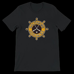 May include: Black t-shirt featuring a yellow and white logo of a ship's wheel with the words "Baltimore Skipjacks" and crossed hockey sticks. The logo is circular and has a vintage design.