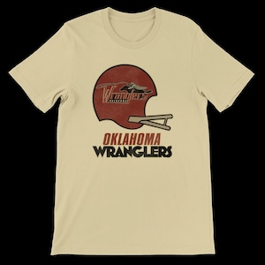 Op de afbeelding: Lichtbeige t-shirt met een bordeauxrood footballhelm-ontwerp en de tekst "Wranglers" en "Oklahoma Wranglers" in zwart en rood. Het shirt heeft korte mouwen en een ronde hals.