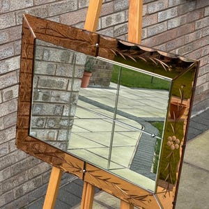 May include: A vintage beveled mirror with a decorative etched floral design in a warm brown tone. The mirror is framed in a beveled glass frame and is standing on a wooden easel.