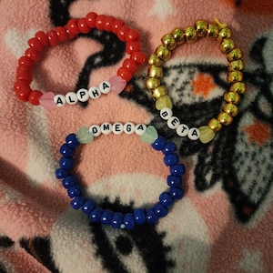 May include: Three friendship bracelets with the words "ALPHA", "OMEGA", and "BETA" spelled out in white beads. The bracelets are red, gold, and blue.