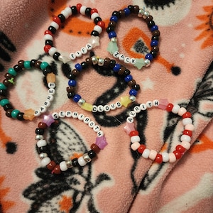 May include: Six colorful beaded bracelets with star charms and names spelled out in white letters. The names are Tifa, Zack, Barrett, Sephiroth, Cloud, and Aerith.