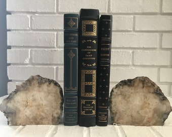 May include: Three black hardback books with gold lettering and decorative designs on the spines. The books are titled "The Sound and the Fury", "The Confessions of Saint Augustine", and "Faust Goethe". The books are being held up by two natural stone bookends.
