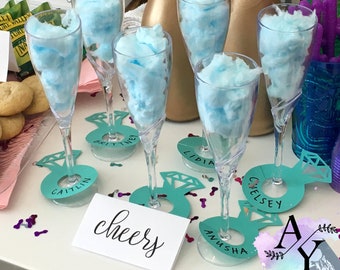 May include: A table setting for a party with six champagne flutes filled with blue cotton candy. Each flute has a teal paper ring with a diamond cutout and a name written on it. The names are Caitlin,  Britney,  Lidia,  Chelsea,  Anusha, and a name that is partially obscured. A sign on the table reads "cheers".