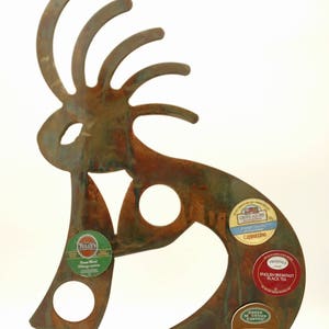 May include: A rusty metal Kokopelli figurine with a round base. The figurine is holding five coffee pods, including English Breakfast Black Tea, Hazelnut Decaf, Milk Chocolate Hot Cocoa, French Vanilla Cappuccino, and House Blend.