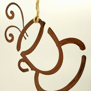 May include: A rustic brown metal ornament shaped like a coffee cup with steam rising from the top. The ornament is hanging from a twine string.