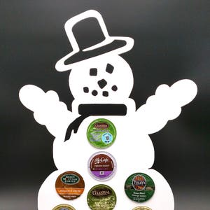May include: A white snowman-shaped coffee pod holder with a black top hat and scarf. It holds various coffee and hot cocoa pods, including brands like Green Mountain Coffee, McCafe, and Grove Square. The pods are in various colors.