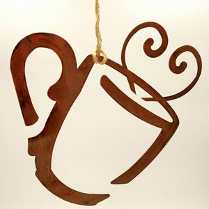 May include: A rustic brown metal ornament shaped like a coffee mug with a swirl of steam coming from the top. The ornament is hanging from a twine string.