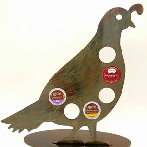 May include: A metal quail-shaped stand with five holes for holding coffee pods. The stand is painted in a rustic brown and green color. The stand has a round base and is decorated with three coffee pod lids, including McCafe French Roast, McCafe Breakfast Blend, and Twinings English Breakfast Black Tea.