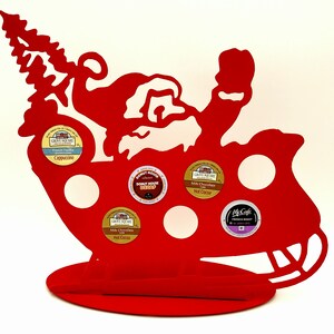 May include: Red metal Santa Claus-shaped coffee pod holder with six slots for coffee pods. The holder has a base and a stand. The coffee pods are labeled with the flavors: Cappuccino, Milk Chocolate Hot Cocoa, Donut House Decaf, Milk Chocolate Hot Cocoa, and French Roast.