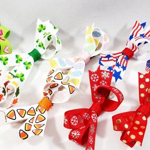 May include: A set of 7 colorful hair bows with various patterns, including hearts, shamrocks, candy corn, snowflakes, and stars. The bows are made of ribbon and are attached to alligator clips.