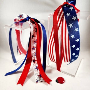 May include: Two sets of red, white, and blue ribbon streamers tied together. The streamers are on a clear acrylic display stand. A small red ladybug is in the foreground.