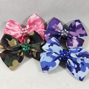 May include: Four hair bows made of ribbed fabric with a camouflage pattern in pink, purple, green, and blue. Each bow features a central cluster of small, sparkling rhinestones that match the color of the camouflage print on the fabric.