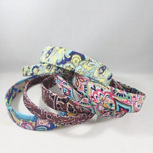 May include: A collection of four fabric-covered headbands featuring various colorful patterns. One headband has a yellow and blue paisley print, another displays a vibrant pink and teal floral design, while two others feature brown and blue paisley motifs.